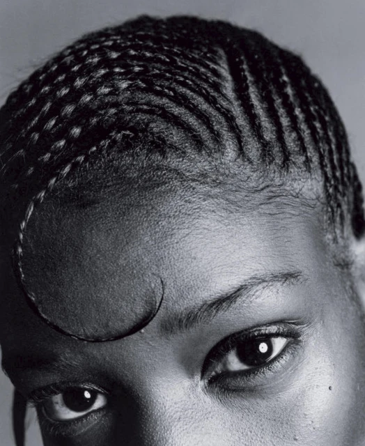 Close-up studio shot of a Black adult female with braided hair and distinct swooped forelock, eyes looking directly at the camera in monochrome.