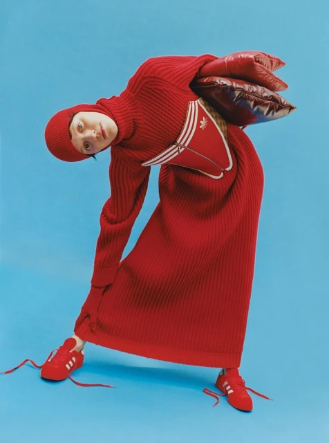 A fashion-forward photo featuring a woman in a full-length red knitted dress and matching beanie, paired with red Adidas sneakers against a blue backdrop.