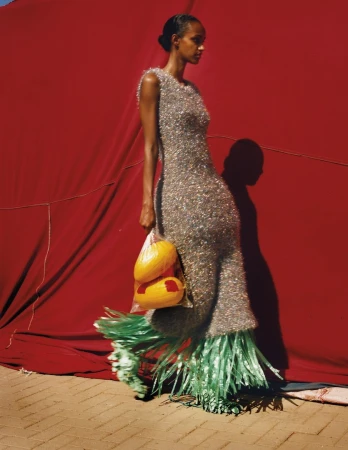 Adult female in a textured gown poses in front of a red curtain holding produce in a bag for a fashion editorial shot.