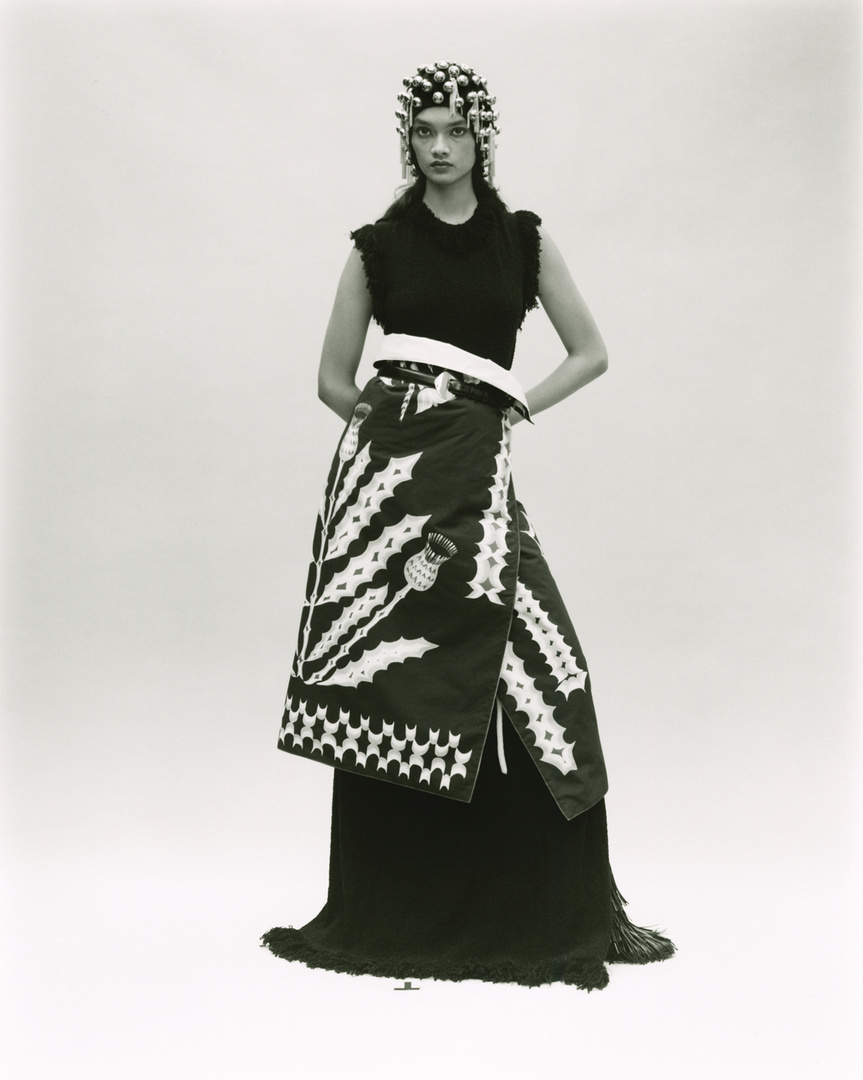 Monochrome fashion portrait of a female model with an elaborate headpiece, sleeveless top, and wrap skirt with thistle design.