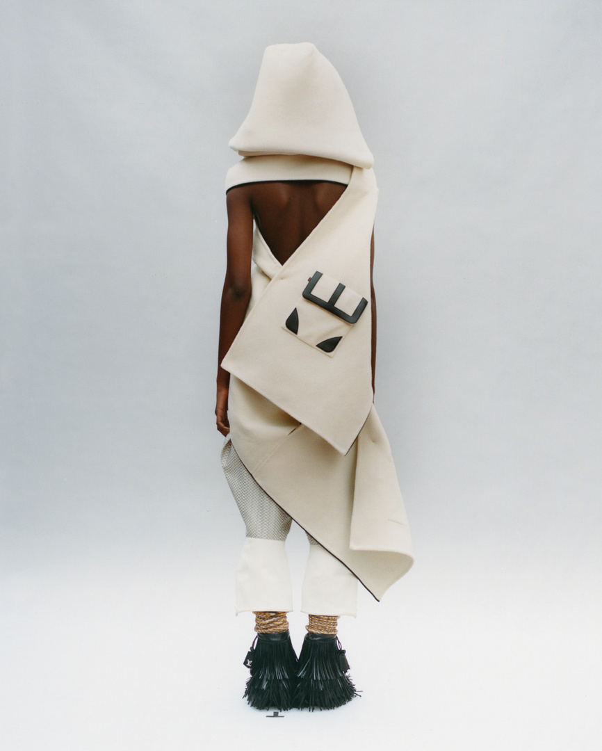 A person wearing an avant-garde hooded garment with a satchel and decorative black fringe shoes, standing in a white photo studio.