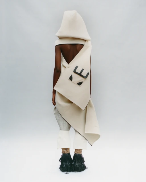 A person wearing an avant-garde hooded garment with a satchel and decorative black fringe shoes, standing in a white photo studio.