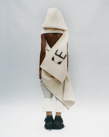 A person wearing an avant-garde hooded garment with a satchel and decorative black fringe shoes, standing in a white photo studio.