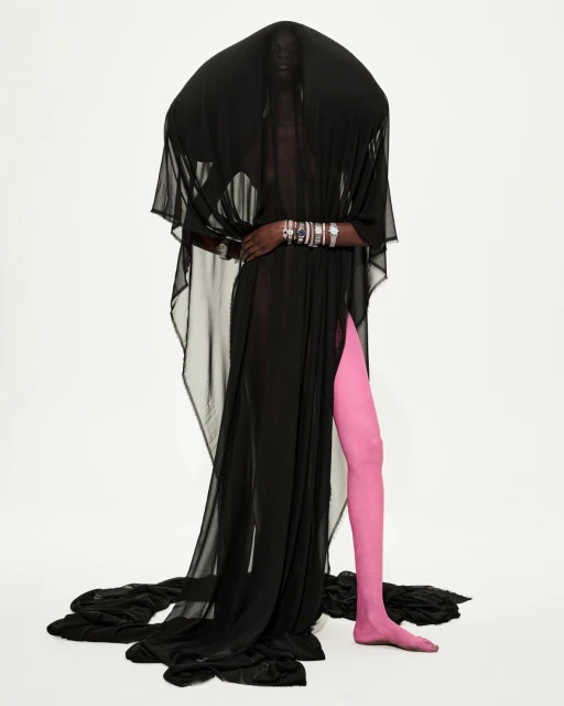 A person draped in flowing black fabric, posing against a white backdrop with pink leggings, adorned with multiple silver bracelets.