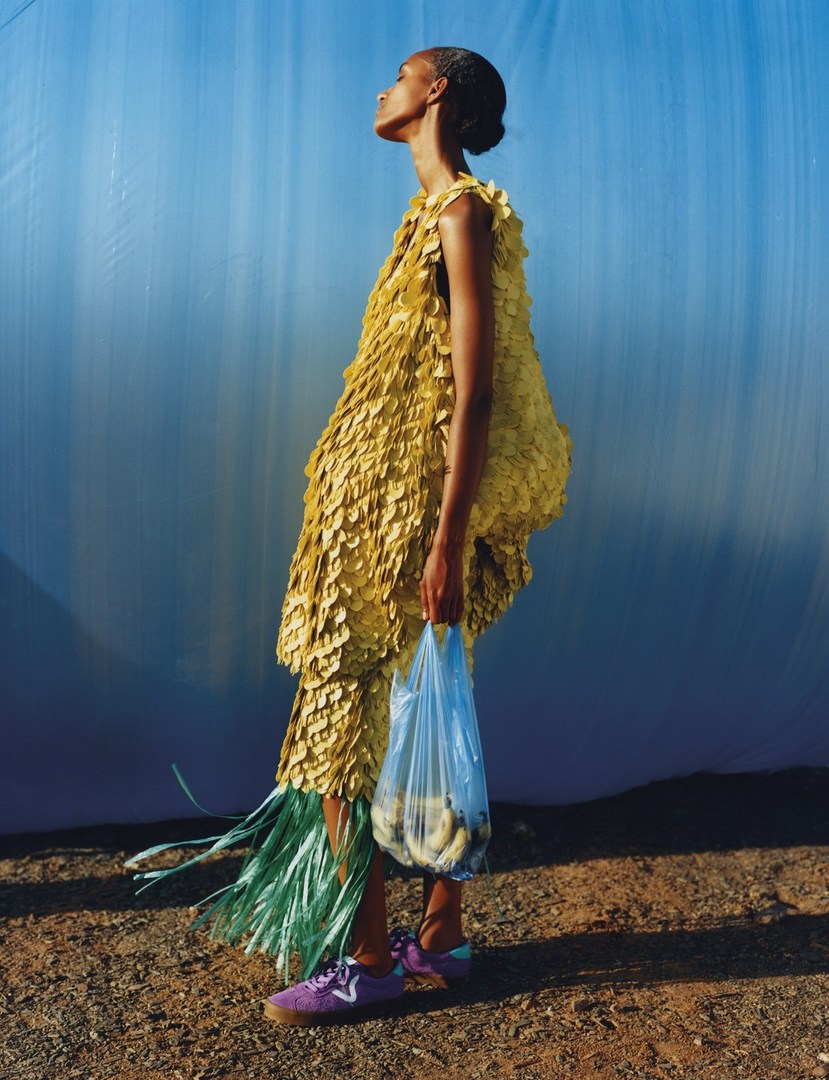 Fashion editorial shot of a Black female model in a yellow dress holding a plastic bag, set against a vibrant blue backdrop and brown ground.