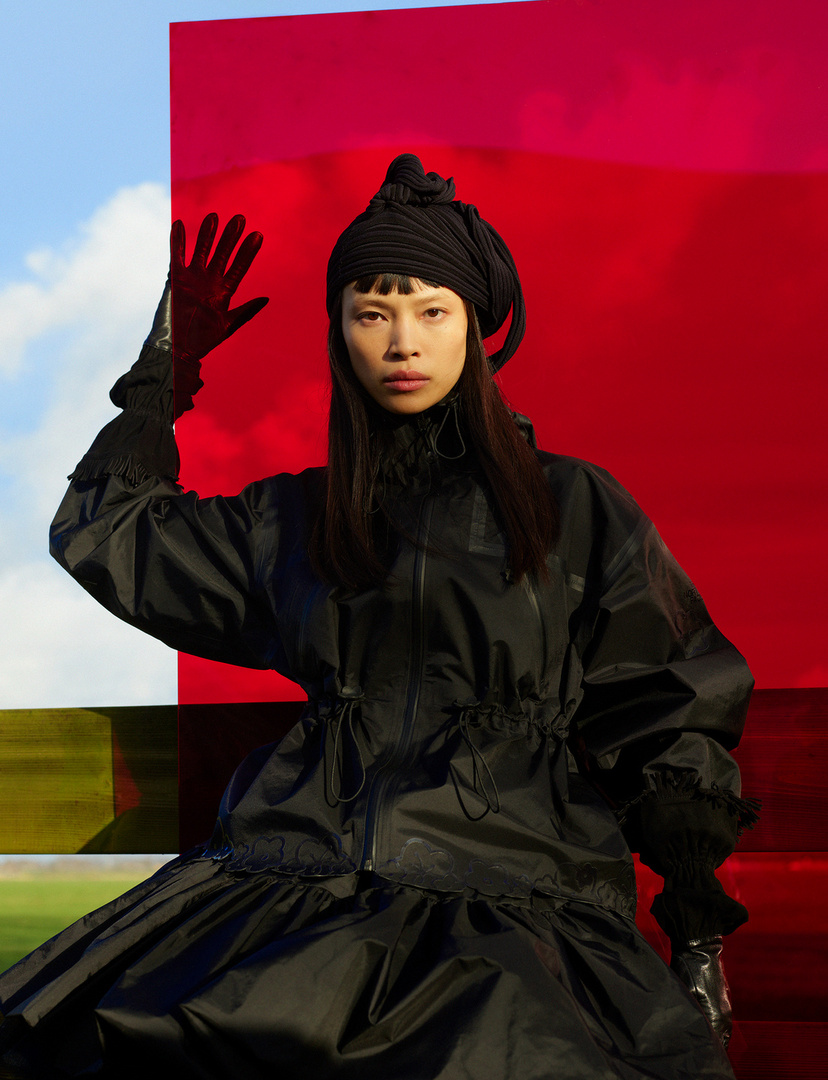 A woman in a black outfit with a head wrap poses against a red panel with blue sky and green field reflections creating a striking contrast.