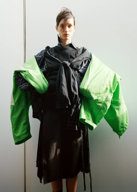Fashion shot of a slender person with short dark hair wearing an assemblage of clothing including a black dress and green jacket.