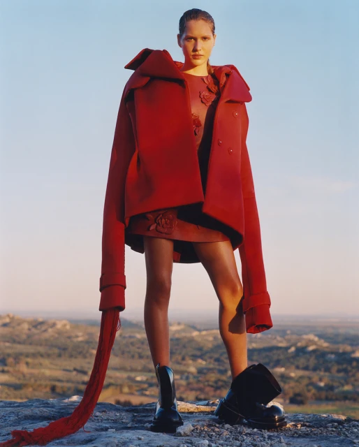 A fashion model in a red oversized coat and leather dress stands on a rocky surface against a pale blue sky in a high-fashion editorial shot.