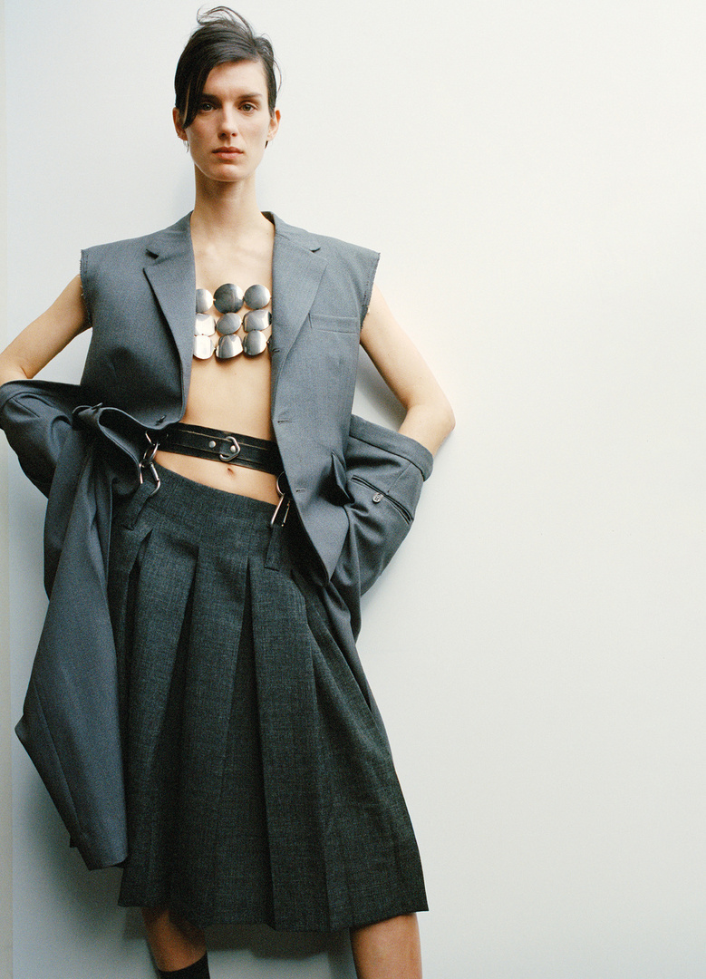 Woman poses in grey sleeveless blazer and pleated skirt with silver body jewelry in studio lighting against a white wall background.
