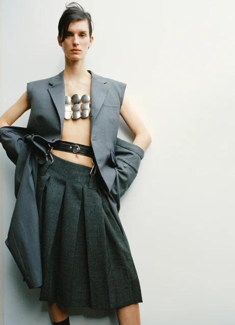 Woman poses in grey sleeveless blazer and pleated skirt with silver body jewelry in studio lighting against a white wall background.