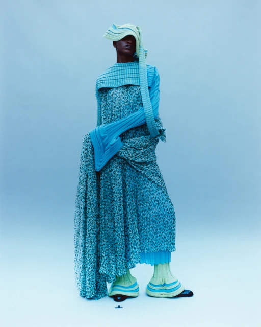 Fashion model in an abstract pose wearing a light blue dress and a matching headpiece, in a studio with a light blue background.