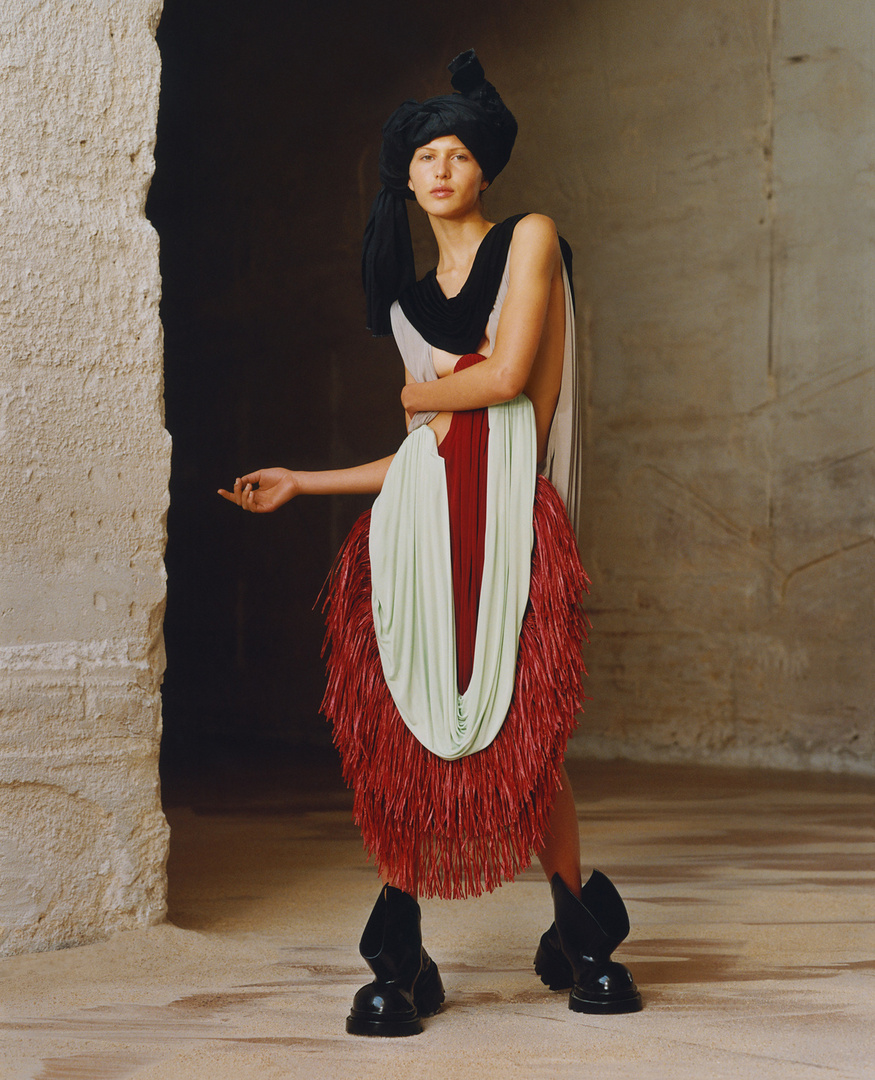 Fashionable woman in abstract outfit featuring black headwrap, light green, red and black color blocks in a cracked-wall setting.
