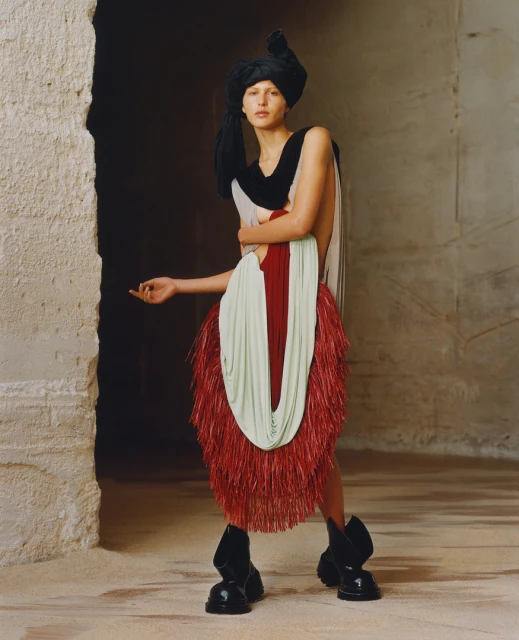 Fashionable woman in abstract outfit featuring black headwrap, light green, red and black color blocks in a cracked-wall setting.