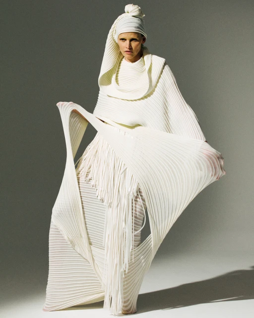 A single fair-skinned adult woman in a light studio wears a light, intricately pleated dress and head covering with an elaborate design.
