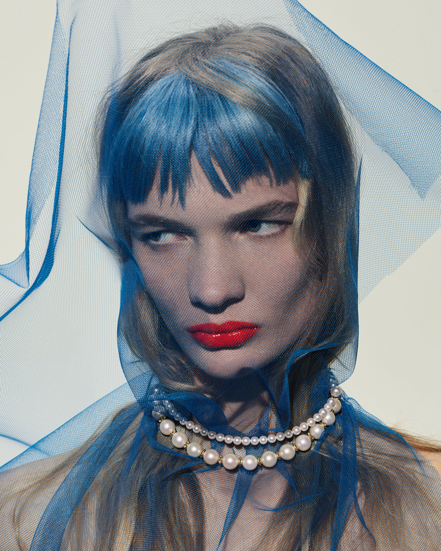 Portrait of a woman with blue-streaked hair, red lipstick, and a pearl necklace, partially veiled in blue netting against a white background.