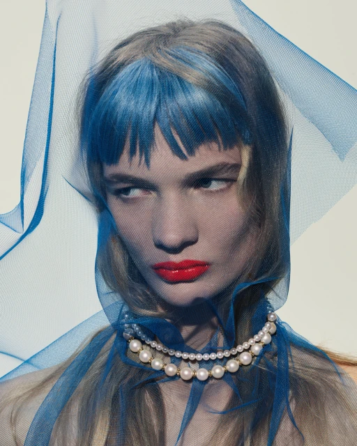 Portrait of a woman with blue-streaked hair, red lipstick, and a pearl necklace, partially veiled in blue netting against a white background.