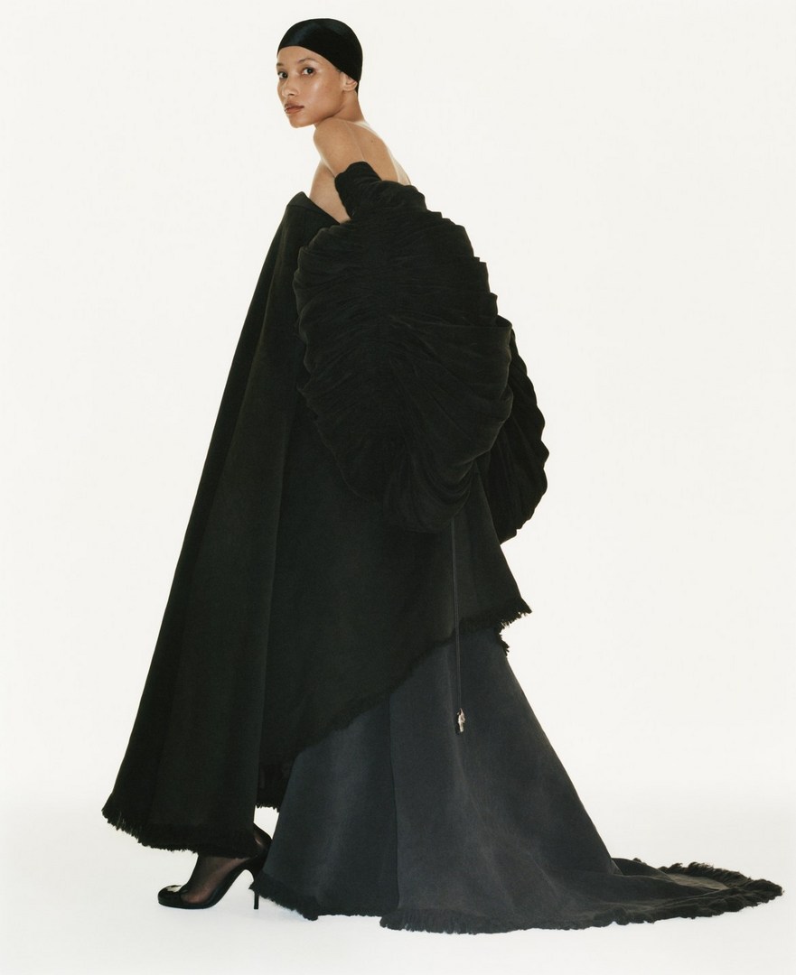 Elegant woman in off-the-shoulder black couture gown and head covering poses in a fashion studio with white backdrop and soft, diffused light.