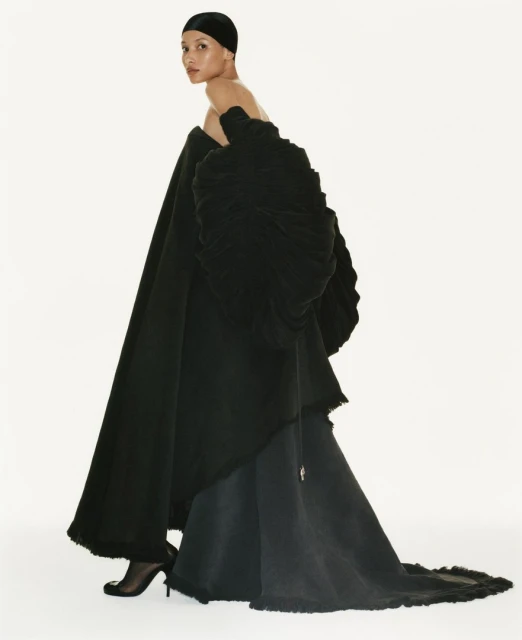 Elegant woman in off-the-shoulder black couture gown and head covering poses in a fashion studio with white backdrop and soft, diffused light.