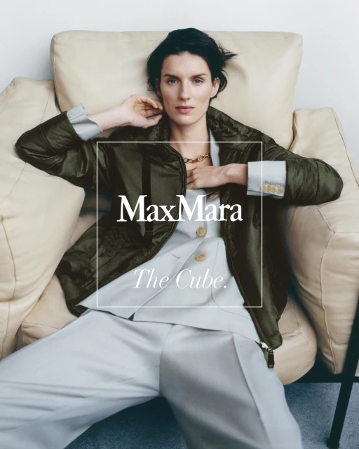 Adult woman in a green puffer jacket and light trousers reclines on a cream-colored armchair in a fashion editorial shot.
