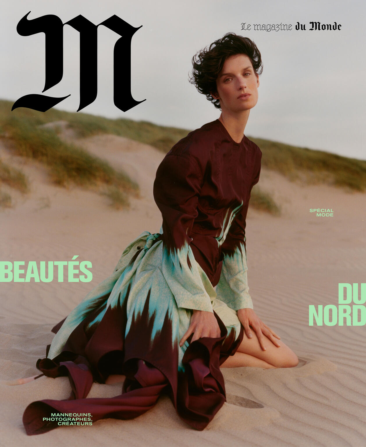 Fashion magazine cover featuring a model in an elegant dress sitting on a sandy dune, with bold typography and a desaturated color palette.