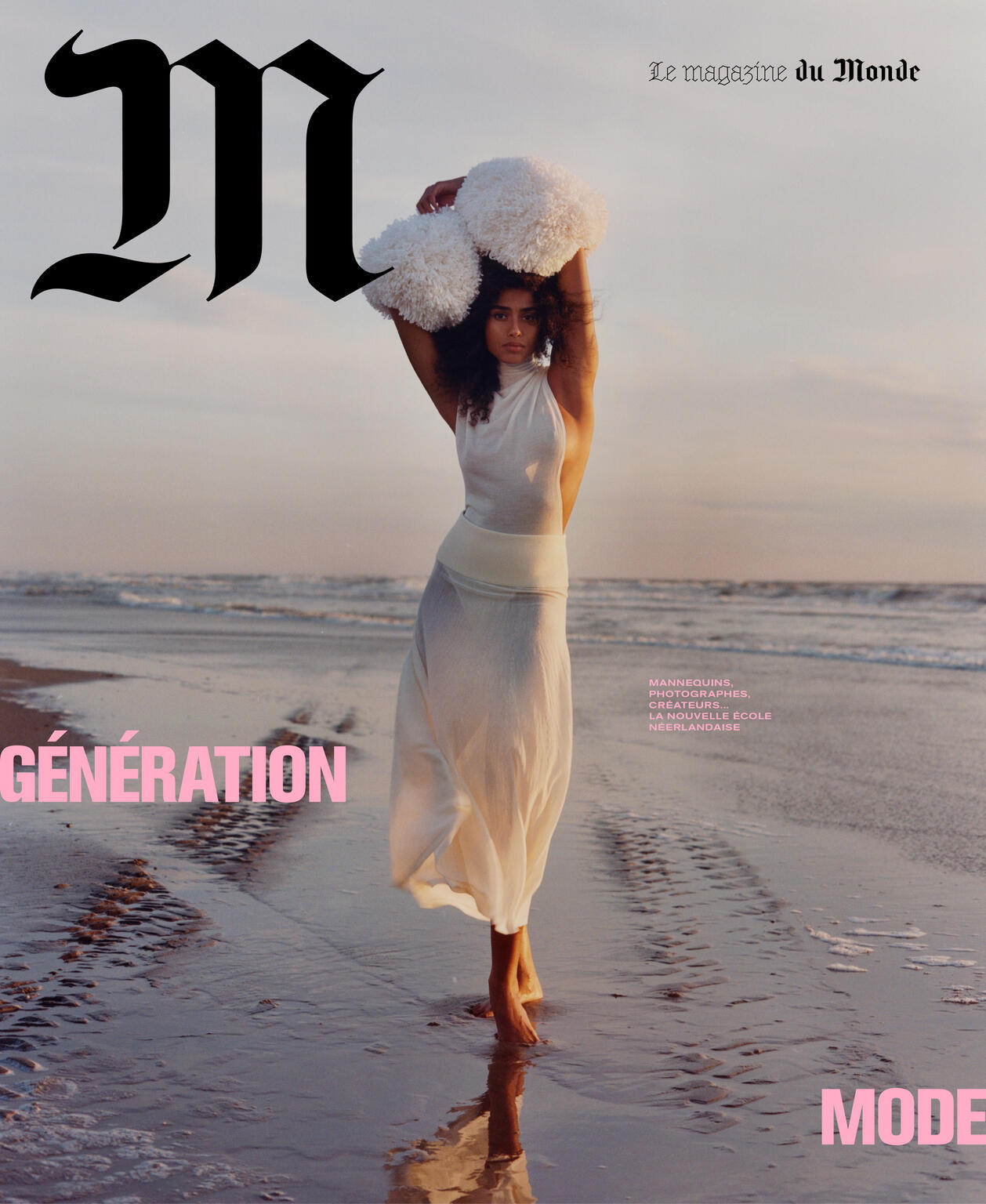 Magazine cover shot of a barefoot adult woman standing on a beach wearing a long white dress, holding white puffy objects above her head.