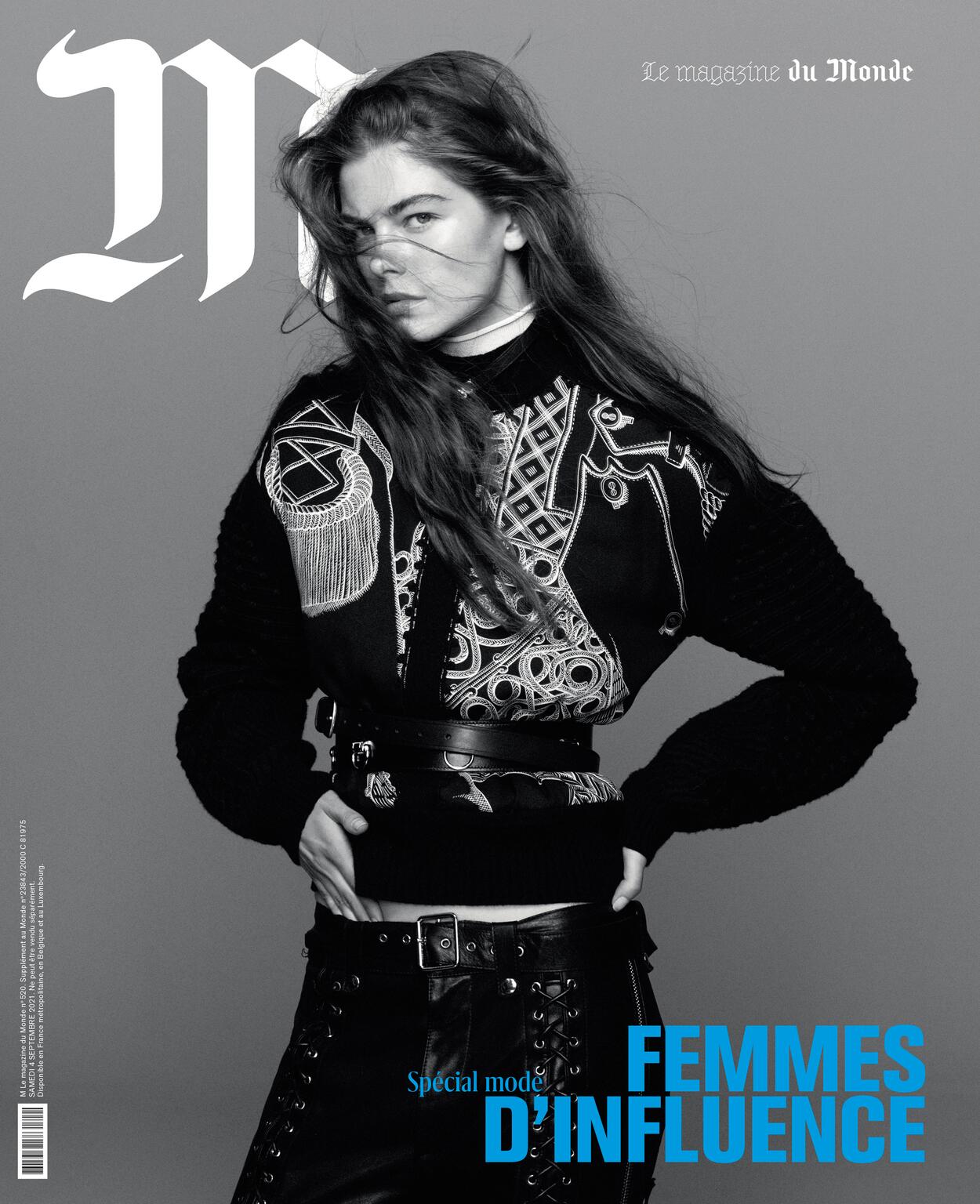Fashion editorial shot of a fair-skinned woman with long hair, wearing a graphic black top, leather pants, and posing against a gray background.