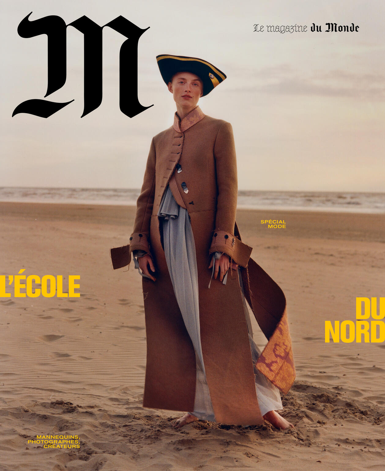 Fashion editorial cover featuring a model on a beach, wearing a brown coat and pirate hat against a blurred horizon backdrop.