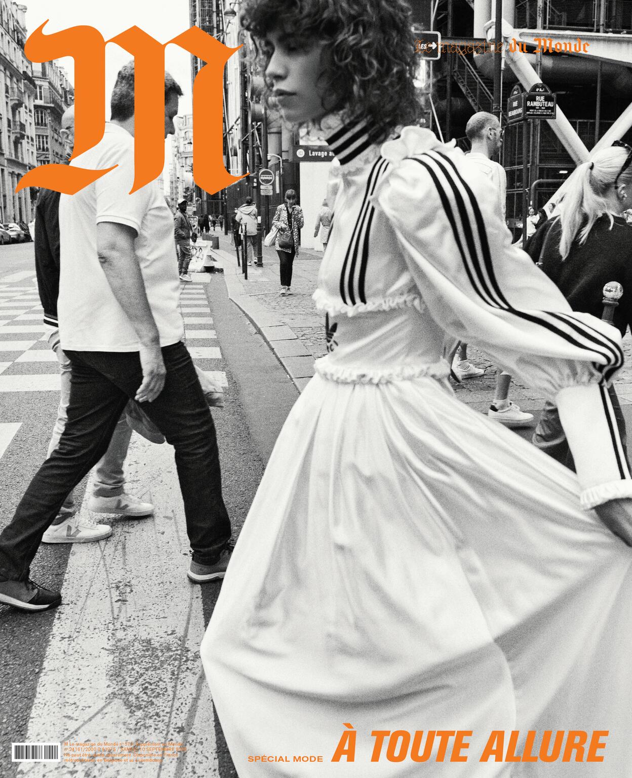 Editorial shot of a curly-haired woman in a white dress and a man in a white shirt crossing a city street with buildings and pedestrians.
