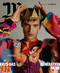 Man wearing a colorful patterned knit sweater and a metallic crown, adjusting it with both hands against a grey background.