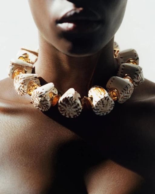 Close-up of a dark-skinned woman wearing a large seashell necklace with gold accents, set against a neutral background in a studio setting.