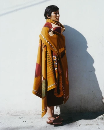 A woman wearing a mustard yellow patterned wrap coat standing against a white wall outdoors in daylight.