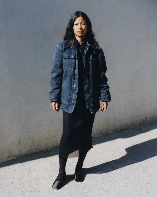 Young woman with long dark hair stands against a concrete wall, wearing a denim jacket, black dress, and black shoes in an outdoor portrait.