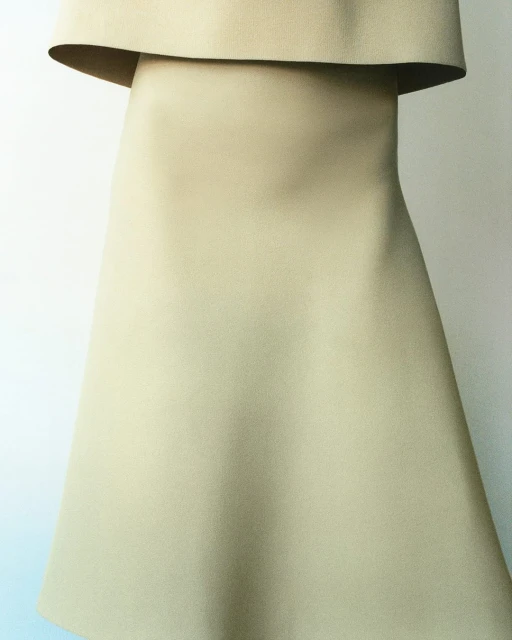 Tan-colored dress with a wide collar, set against a plain white backdrop in a studio-lit fashion shot emphasizing the garment's clean lines.