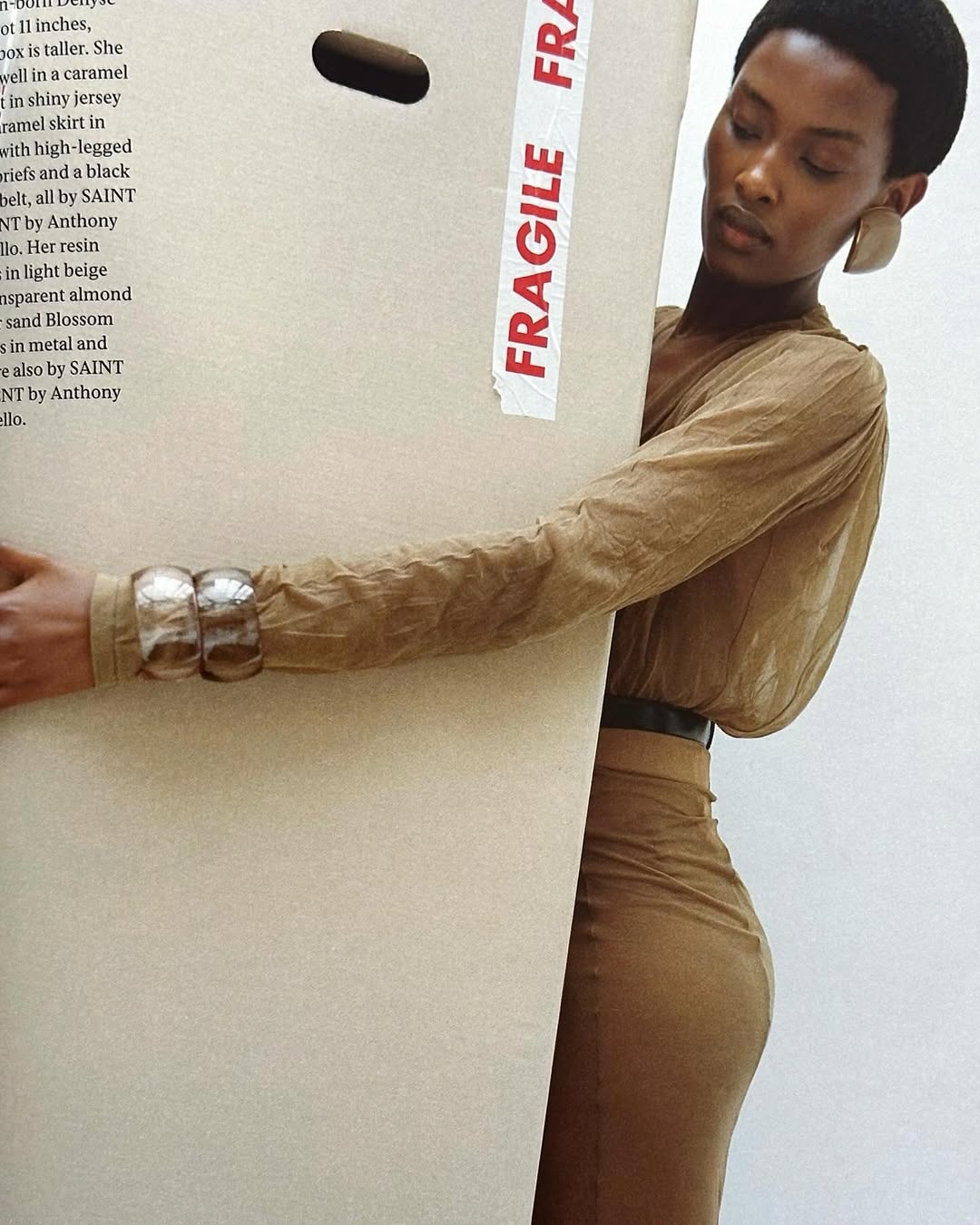 Adult Black woman in a tan dress and jewelry posing with a large carton with 'Fragile' tape, against a neutral white background.