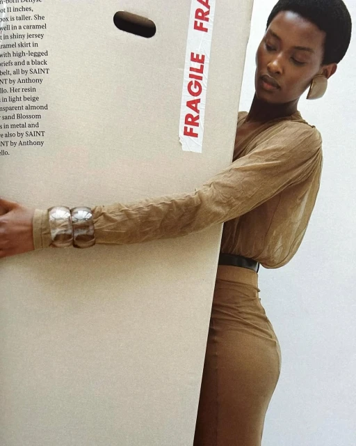 Adult Black woman in a tan dress and jewelry posing with a large carton with 'Fragile' tape, against a neutral white background.