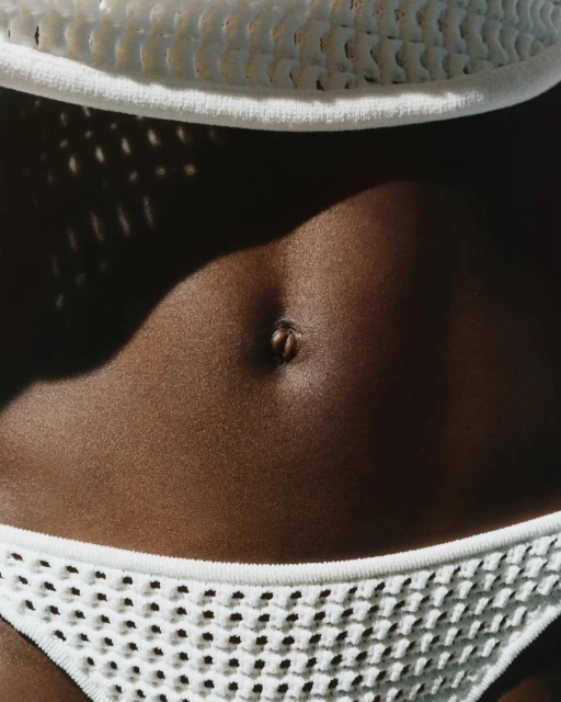 Close-up of a tanned woman's midsection wearing a white crocheted bikini top and bottom with bright, natural light and shadow.