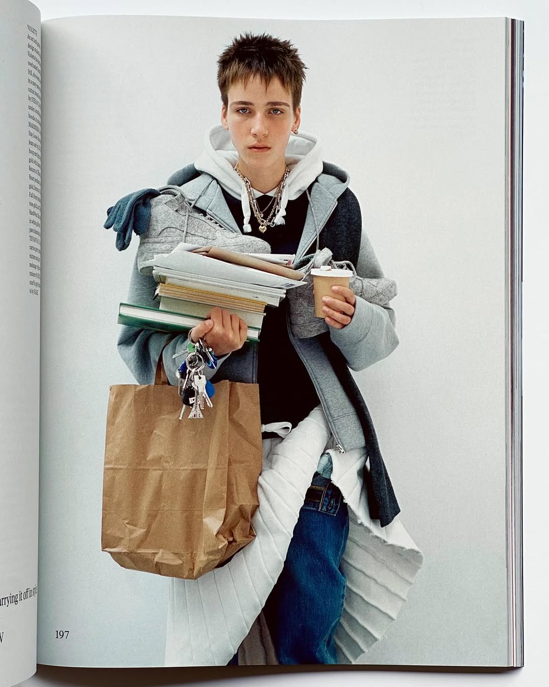 Young adult with short hair in layered clothing, holding books, a paper bag and coffee, against a bright white background.