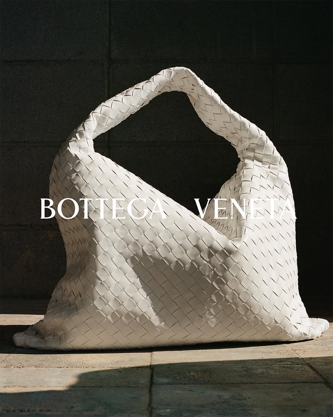 White woven leather handbag by Bottega Veneta displayed indoors on a tiled surface with a dark wall in the background.