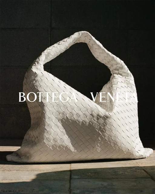White woven leather handbag by Bottega Veneta displayed indoors on a tiled surface with a dark wall in the background.