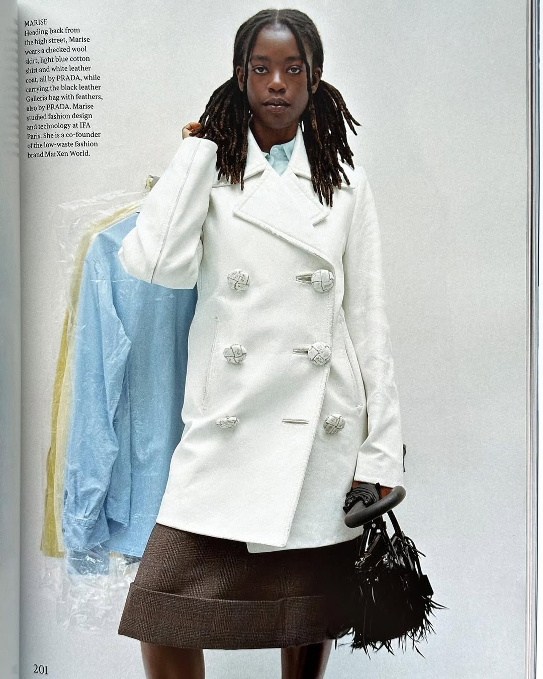Elegant Black woman poses in a white double-breasted coat and brown skirt against a muted studio backdrop, holding a black handbag.