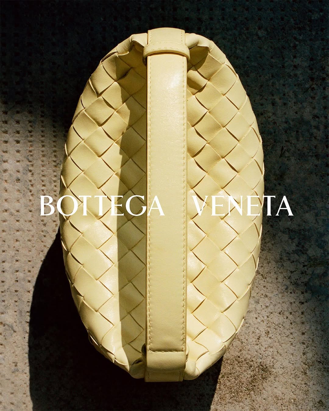 Close-up studio shot of a Bottega Veneta pouch bag with a woven design and a handle in a pale yellow color.
