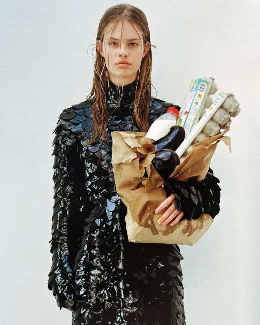 A woman in a dark, scale-like dress stands with a brown paper bag full of groceries against a plain white background, in studio lighting.
