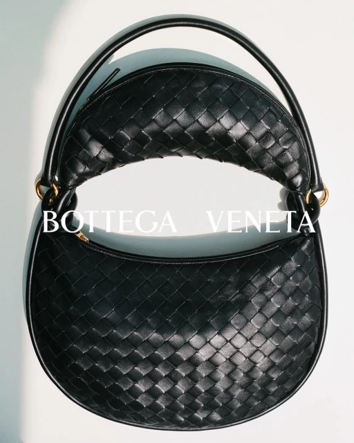 Close-up of a black Bottega Veneta woven leather handbag with a rounded shape and handle, set against a neutral background.