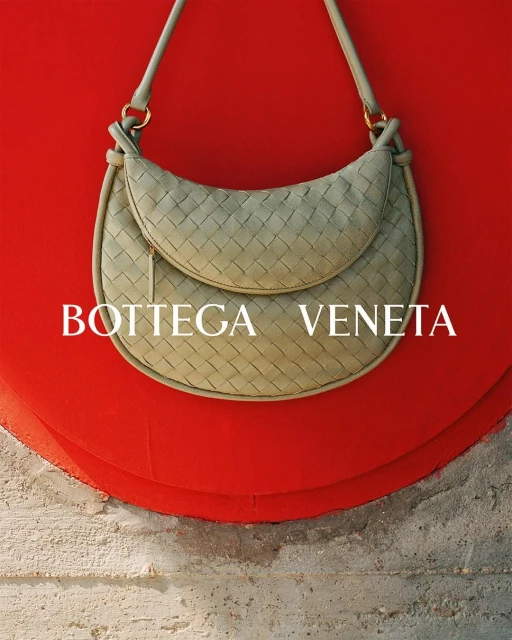 Bottega Veneta woven light green hobo bag with shoulder strap, displayed against red background and textured neutral surface.