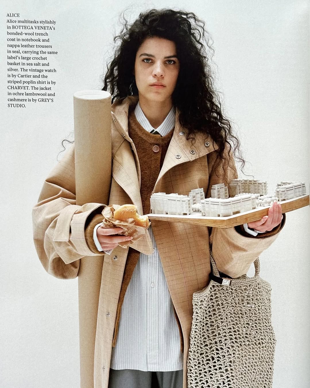 Editorial shot of a woman in a trench coat holding an architectural model, a pastry, and a blueprint, with a tote bag.