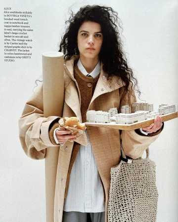 Editorial shot of a woman in a trench coat holding an architectural model, a pastry, and a blueprint, with a tote bag.