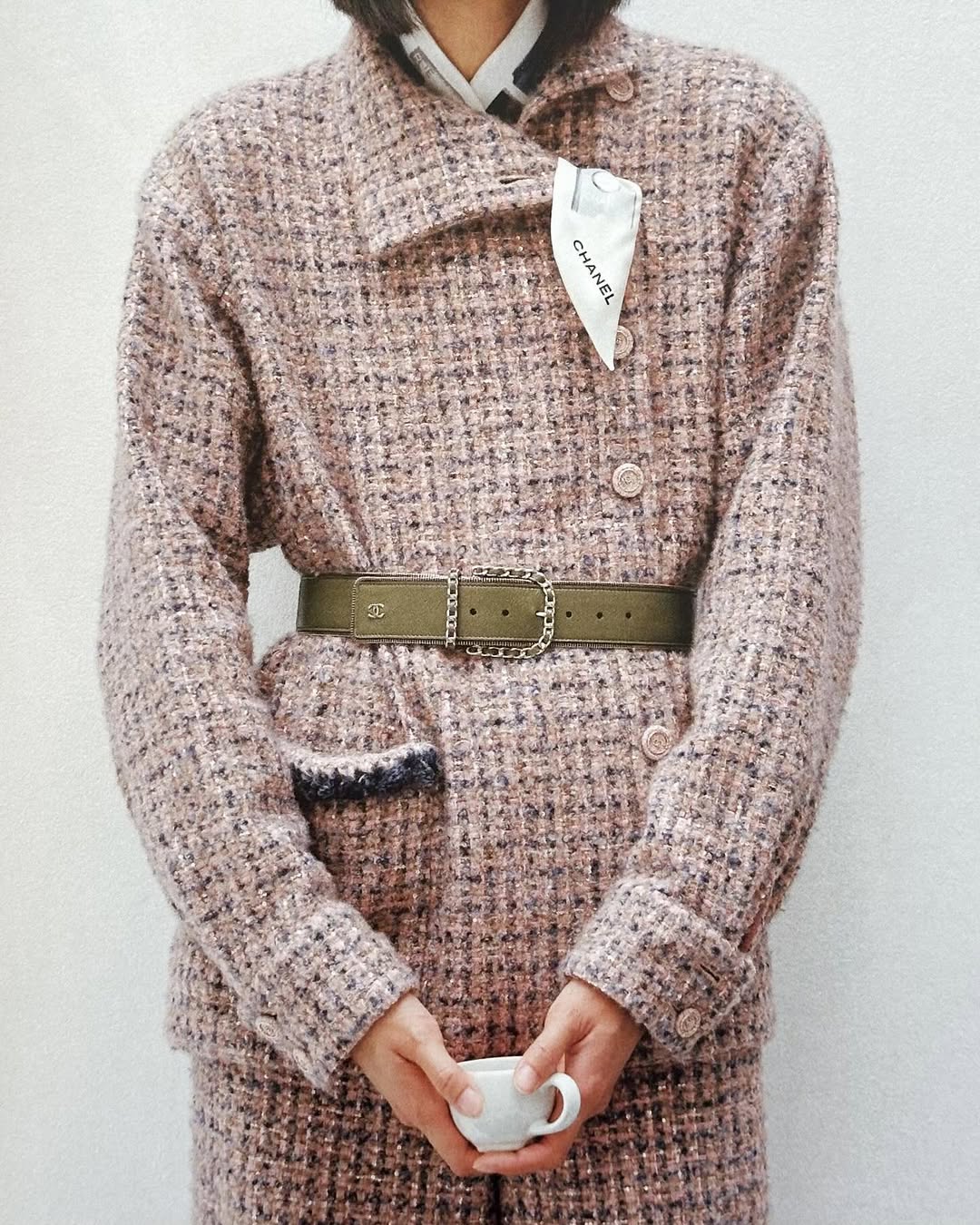 Close-up fashion shot of a person wearing a tweed blazer with a belt, holding a cup in front of a plain white background.