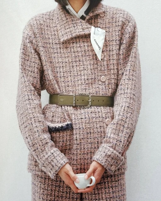 Close-up fashion shot of a person wearing a tweed blazer with a belt, holding a cup in front of a plain white background.