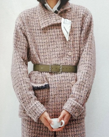 Close-up fashion shot of a person wearing a tweed blazer with a belt, holding a cup in front of a plain white background.