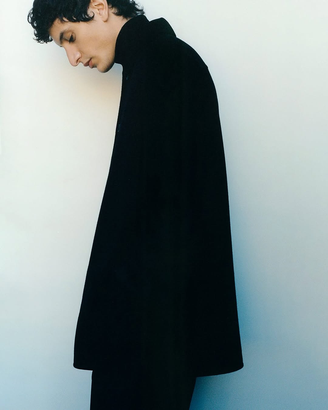 Profile portrait of an adult male model wearing a black turtleneck sweater against a white backdrop, minimalist fashion and studio lighting.